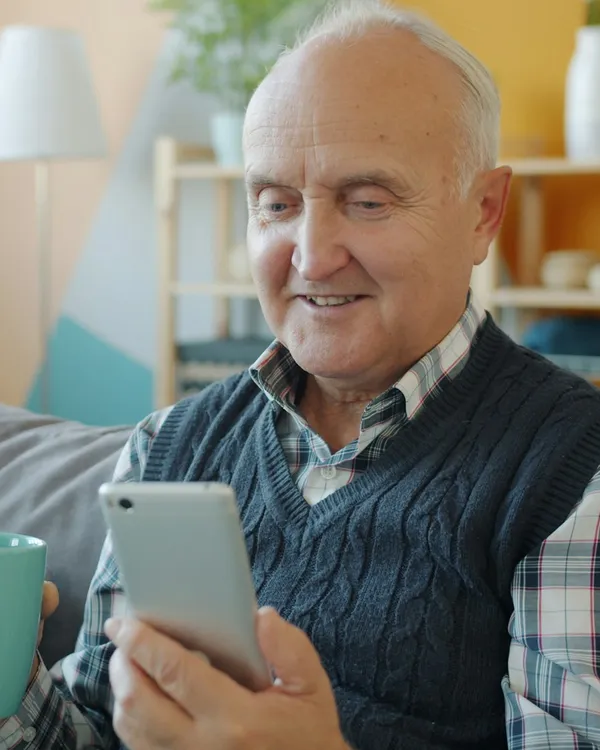 Ein lächelnder älterer Herr mit Pullunder sitzt mit Kaffeetasse auf dem Sofa und spricht mit Anna über sein Handy.