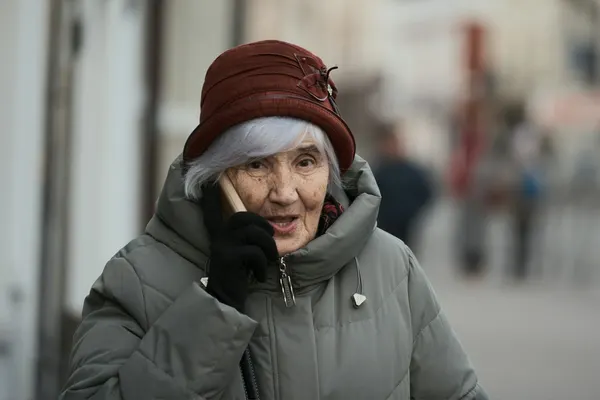Eine ältere Dame mit Hut steht auf einer belebten Straße und telefoniert aufmerksam.